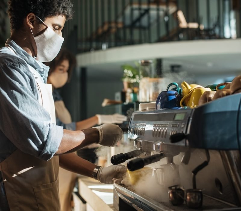 Coffee shop or small business open after lockdown quarantine. Millennial african american barista working with gloves and face mask, make hot drink takeaway on espresso machine in cafe, copy space