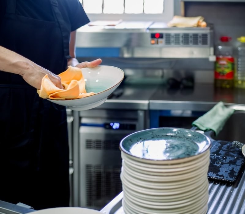 Crop anonymous female chef in black casual clothes with apron washing and wiping ceramic plates stacked on stainless sink at kitchen of restaurant in daylight